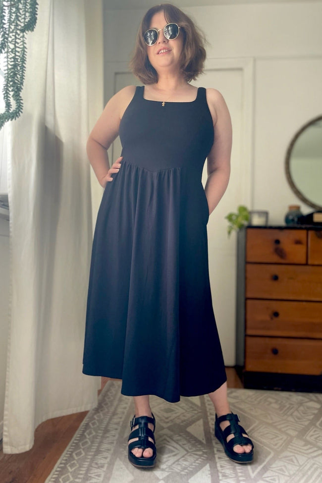 A person wearing a midi length black jersey dress with a square neckline, sleeveless design, and side pockets. The dress features a corset inspired bodice and a center front dip waist.
