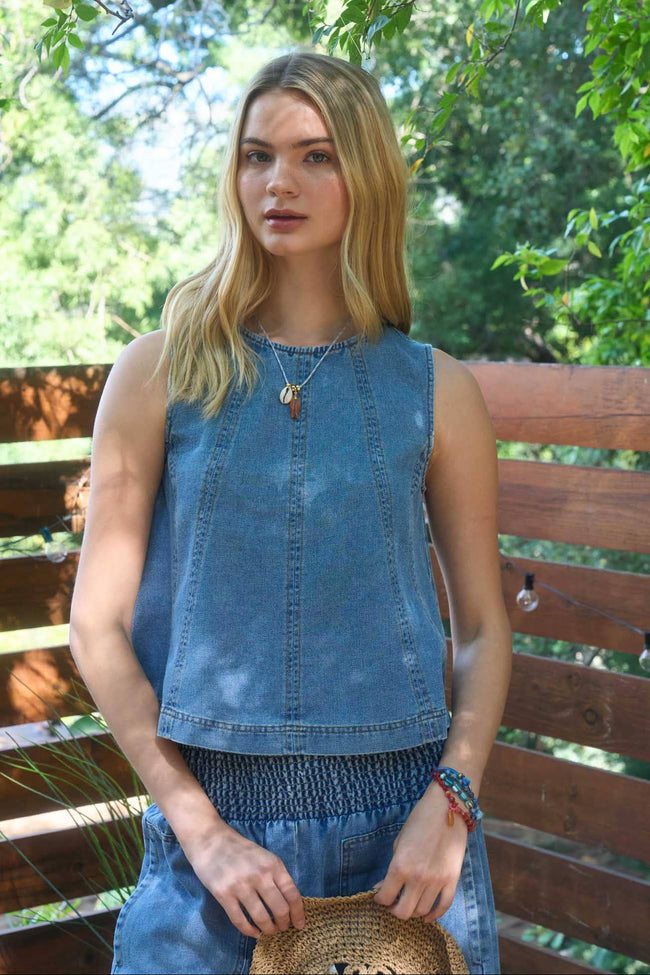 Woman wearing a blue sleeveless top and denim shorts standing outdoors with greenery in the background.