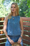 Woman wearing a blue sleeveless top and denim shorts standing outdoors with greenery in the background.