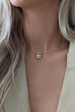 A close-up of a model wearing a delicate gold chain necklace with a small square bezel-set clear gemstone pendant, paired with a beige blazer.