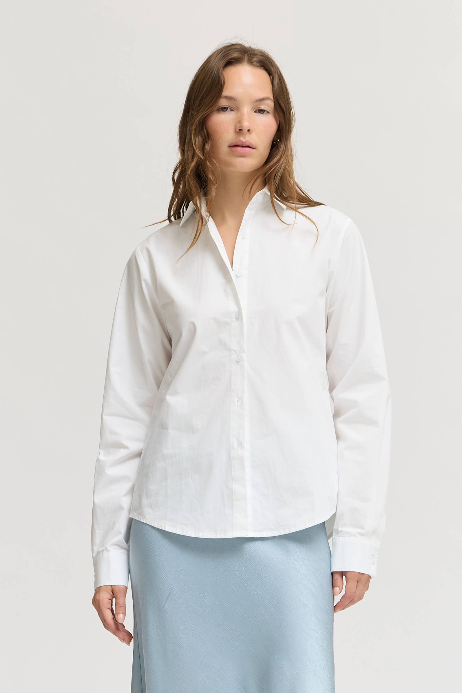 A woman modeling a white long-sleeve button-up shirt with a relaxed fit, paired with a light blue satin skirt, against a plain light gray studio background.