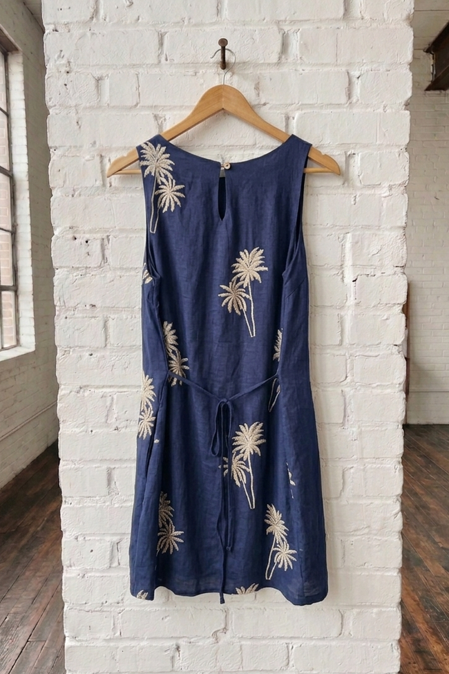 Navy dress with white palm tree embroidered patterns on a wooden hanger against a white brick wall.