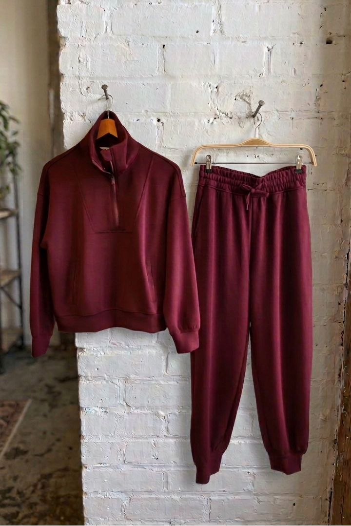 A matching burgundy tracksuit hanging on a rustic white brick wall, featuring a quarter-zip pullover sweatshirt on a wooden hanger and matching jogger sweatpants on a white hanger.