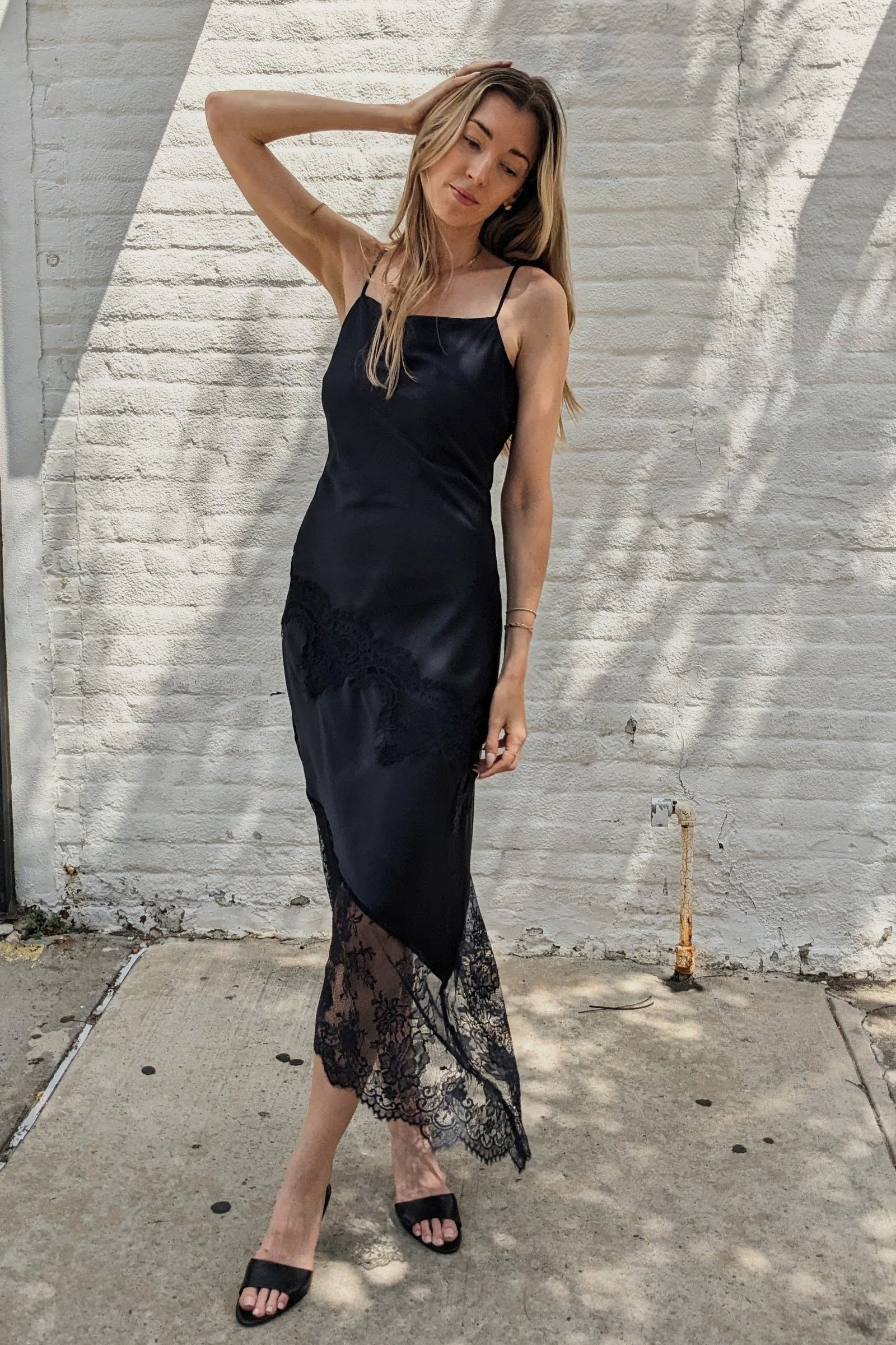 A woman modeling a black satin slip dress with a sheer lace hem, standing outdoors against a white brick wall.