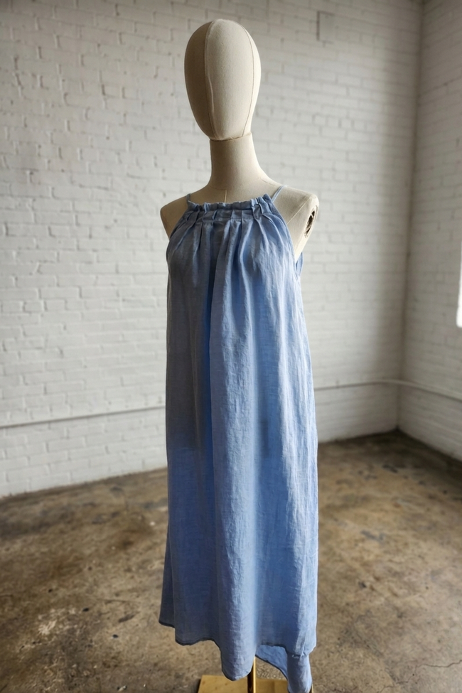 A light blue sleeveless linen dress displayed on a cream mannequin in a bright, white-walled room with a concrete floor.