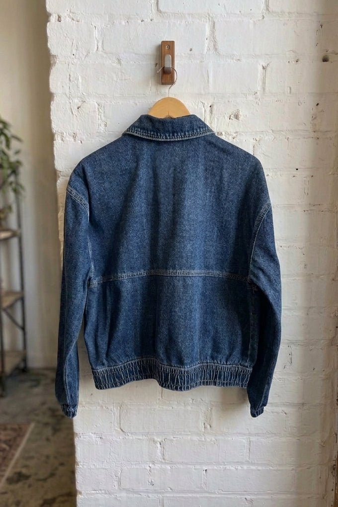 A blue denim jacket hanging on a wooden hanger from a wall hook, shown from the back against a white brick wall in an indoor room setting.