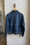 A blue denim jacket hanging on a wooden hanger from a wall hook, shown from the back against a white brick wall in an indoor room setting.
