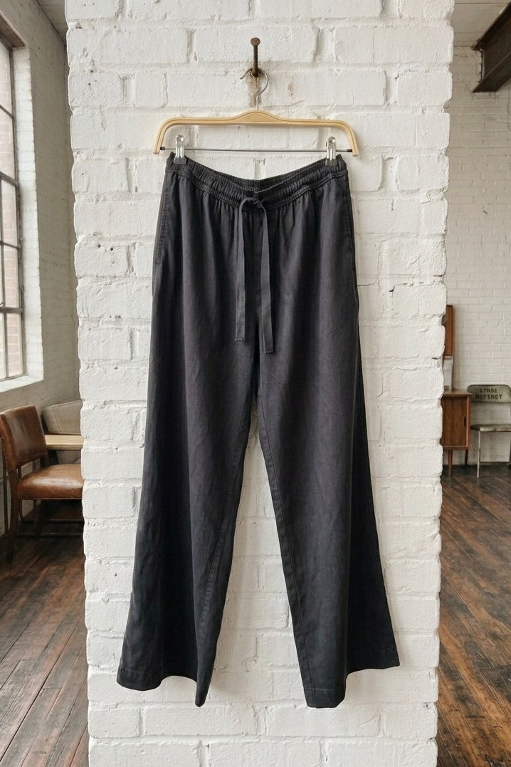 Black wide-leg drawstring pants hanging on a wooden hanger against a white brick wall in a loft-style room with wood floors.