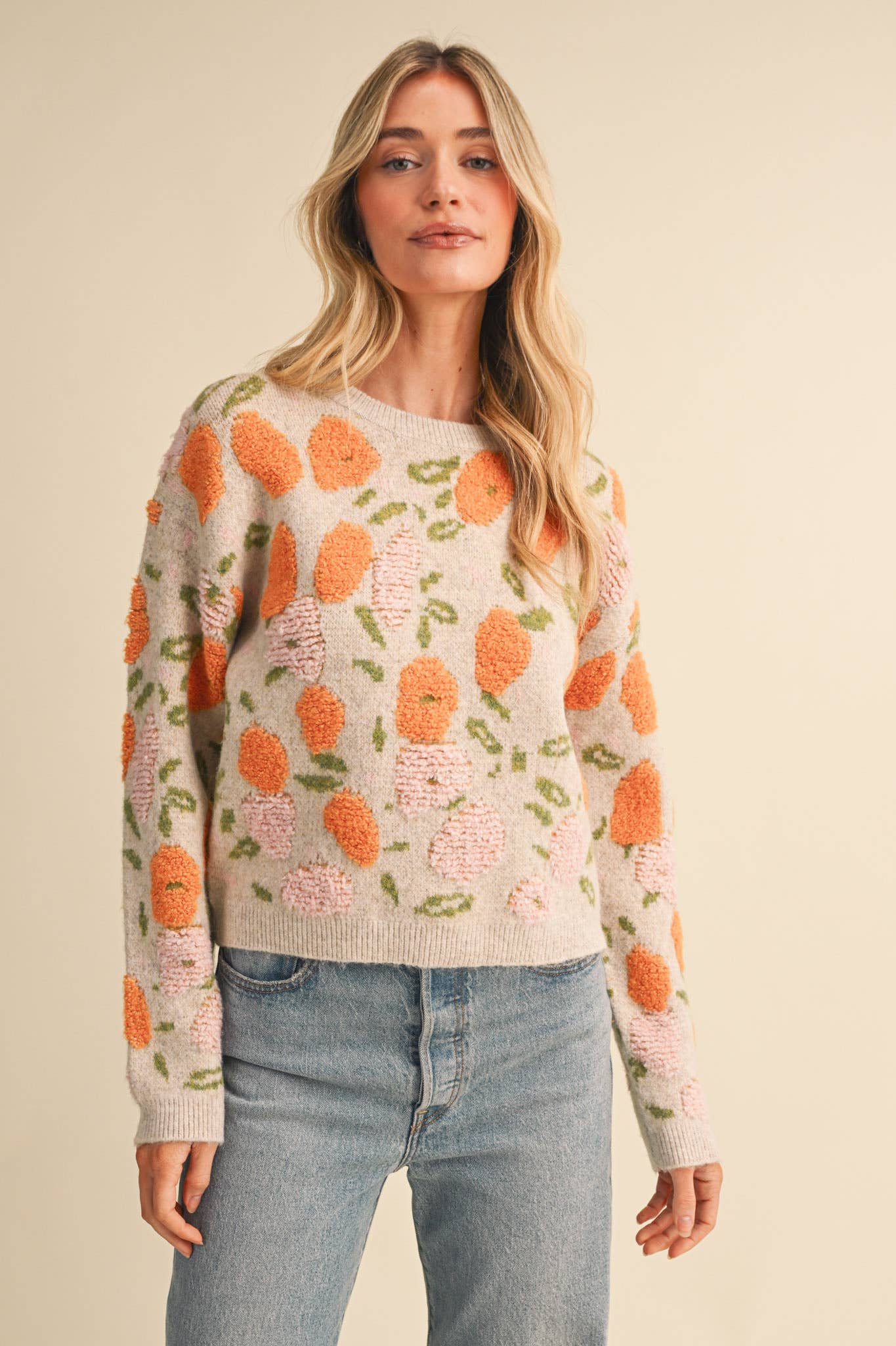 A woman wearing a cream-colored knit sweater with a raised orange and pink floral pattern, paired with light-wash blue jeans, against a solid beige background.