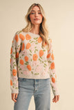 A woman wearing a cream-colored knit sweater with a raised orange and pink floral pattern, paired with light-wash blue jeans, against a solid beige background.