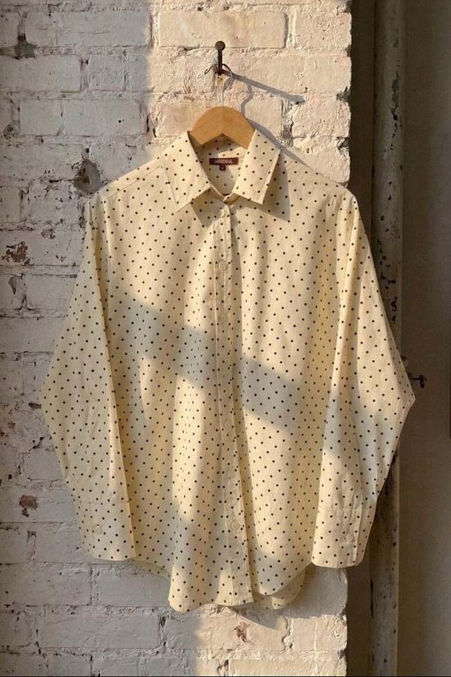 A cream-colored long-sleeve button-up shirt with a small black polka dot pattern, hanging on a wooden hanger from a hook on a white brick wall.