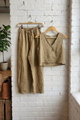 Beige linen pants and crop top hanging on a wooden hanger against a white brick wall.