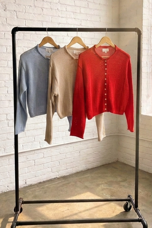 A black metal rolling garment rack holding three long-sleeve knit tops in light blue, beige, and red, set in a bright room with a white brick wall and large window.