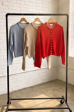 A black metal rolling garment rack holding three long-sleeve knit tops in light blue, beige, and red, set in a bright room with a white brick wall and large window.