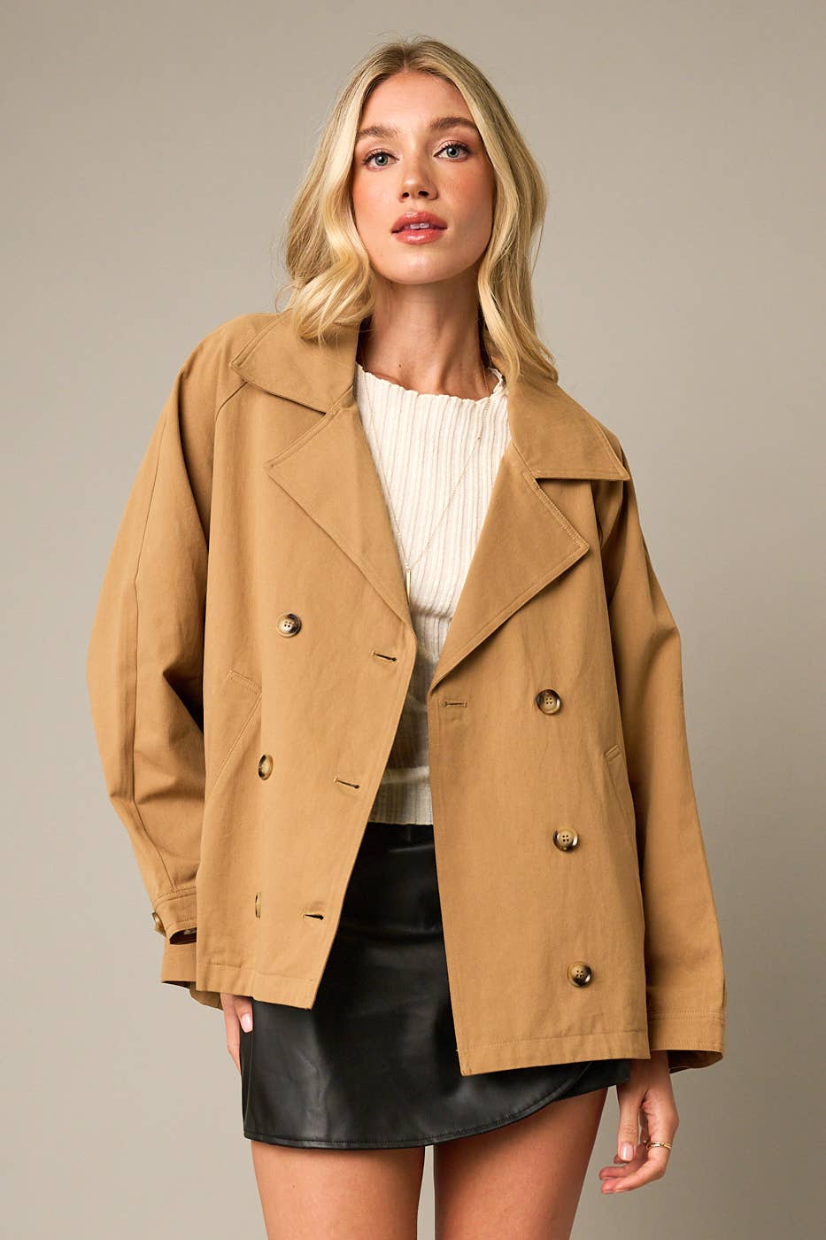 Woman wearing a tan coat over a white sweater and black skirt on a beige background