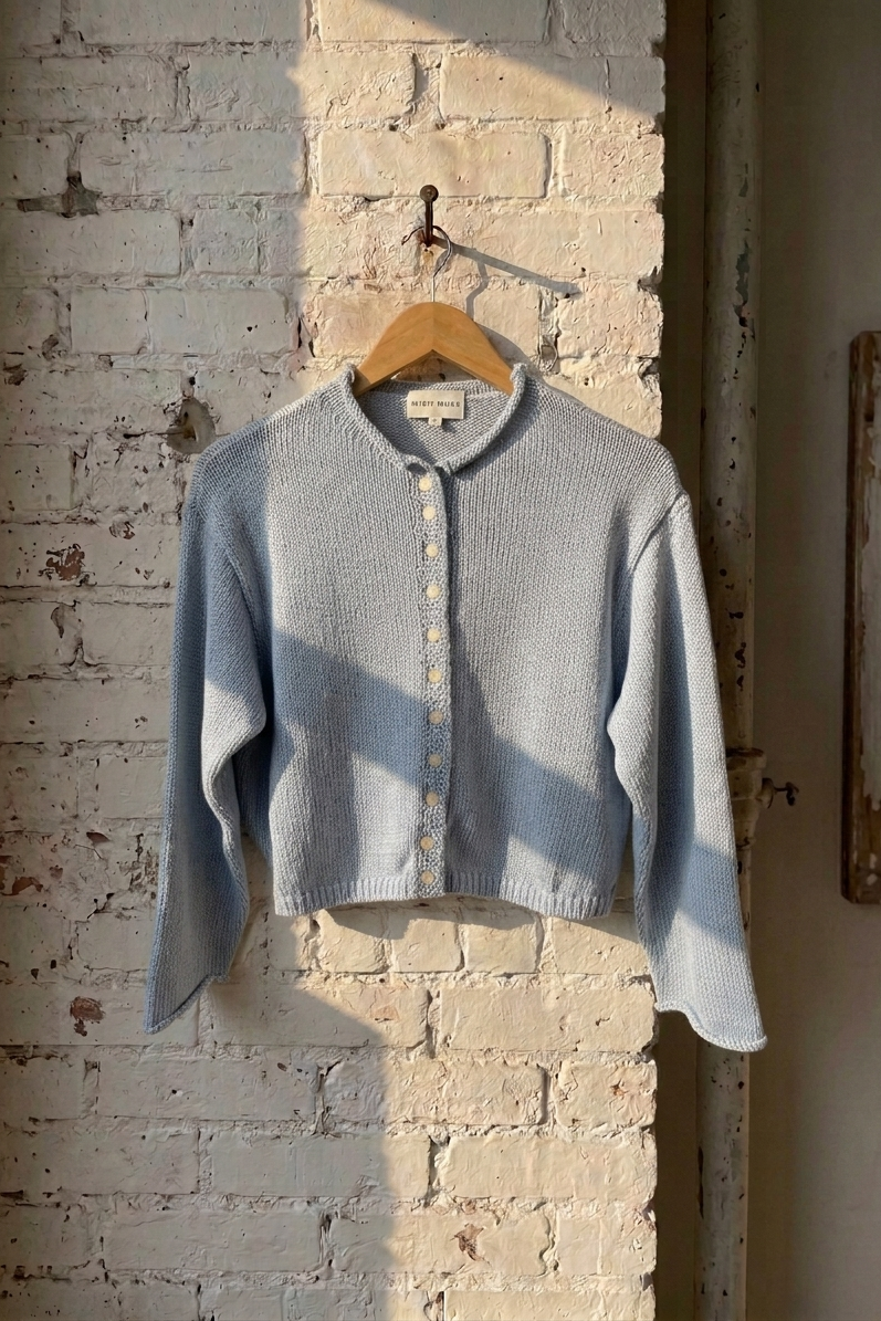 A light blue ribbed knit cardigan sweater with a button-front closure and stand collar, hanging on a wooden hanger against a distressed white brick wall.