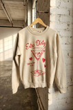 Beige sweatshirt with red graphic and text on a hanger against a brick wall.