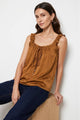 A woman wearing a brown ruffled sleeveless top with a drawstring neckline, paired with dark blue jeans, sitting on a wooden block against a light gray background.