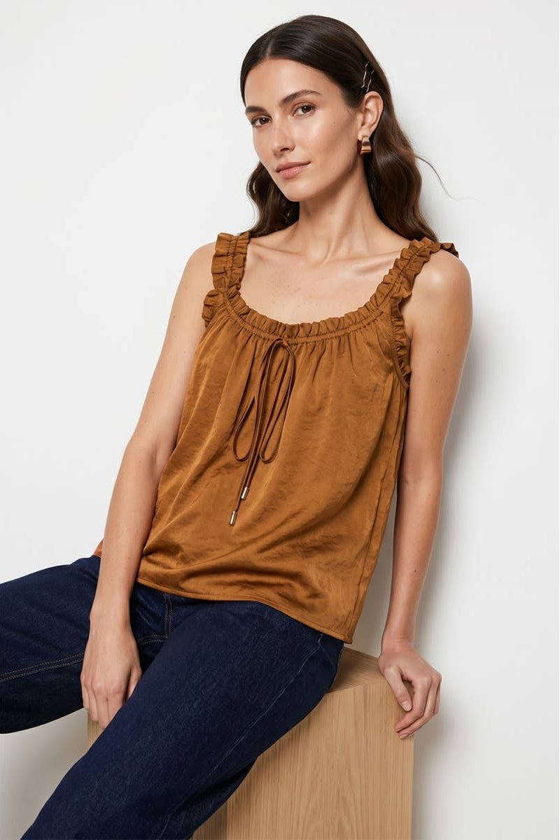 A woman wearing a brown ruffled sleeveless top with a drawstring neckline, paired with dark blue jeans, sitting on a wooden block against a light gray background.