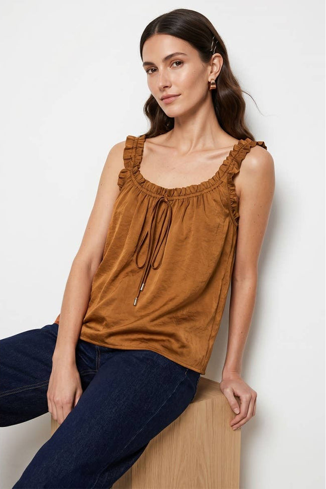 A woman wearing a brown ruffled sleeveless top with a drawstring neckline, paired with dark blue jeans, sitting on a wooden block against a light gray background.