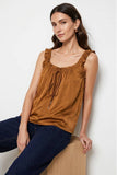 A woman wearing a brown ruffled sleeveless top with a drawstring neckline, paired with dark blue jeans, sitting on a wooden block against a light gray background.