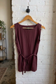 A sleeveless, scoop-neck top in a deep burgundy or plum color, hanging on a wooden hanger against a white-painted brick wall.