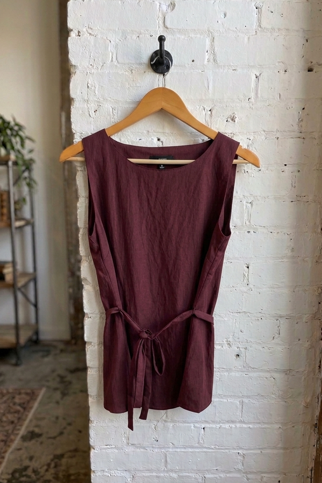 A sleeveless, scoop-neck top in a deep burgundy or plum color, hanging on a wooden hanger against a white-painted brick wall.