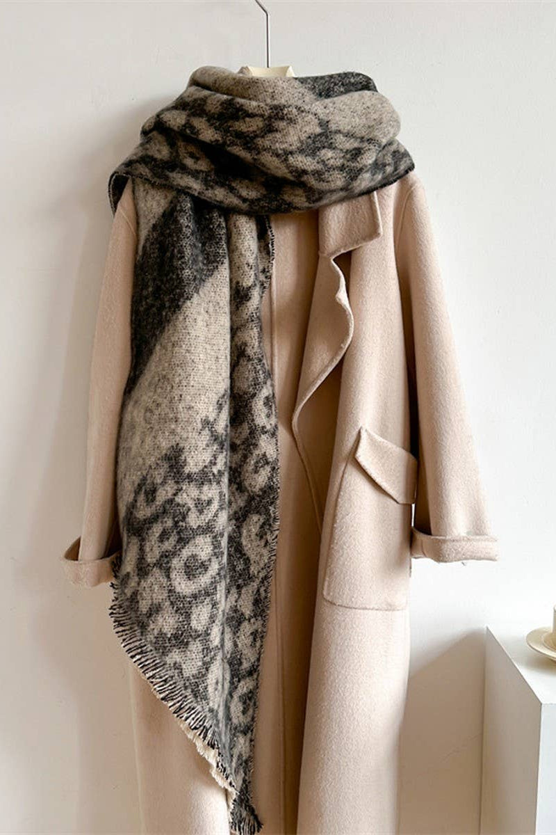 A beige wool coat hanging on a hanger with a large patterned scarf draped over it, set against a plain light wall with a small table and cup at the side.