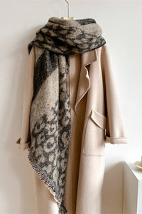 A beige wool coat hanging on a hanger with a large patterned scarf draped over it, set against a plain light wall with a small table and cup at the side.