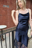 A woman wearing a navy blue satin slip dress with a lace hem, standing on a balcony with a brick wall background.