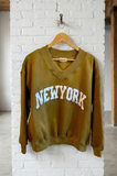 Green sweatshirt with 'New York' text on a wooden hanger against a white brick wall.