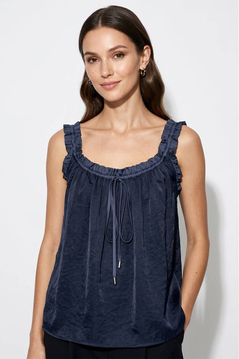 A woman wearing a navy blue sleeveless top with ruffled trim and a drawstring neckline, photographed against a light gray studio background.