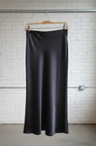 Black satin skirt on a hanger against a white brick wall.