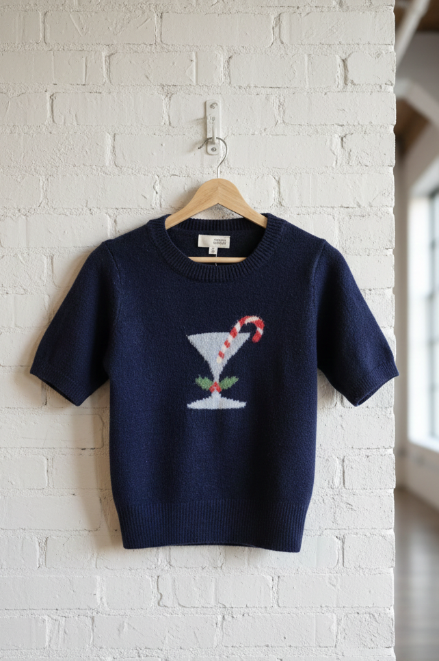 Navy sweater with a martini glass design on a hanger against a white brick wall.