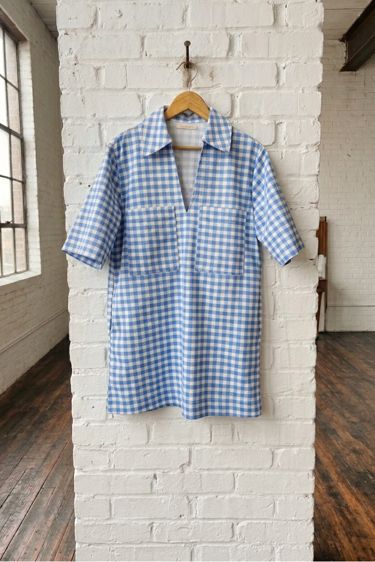 Blue and white checkered dress hanging on a wooden hanger against a white brick wall.