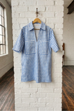 Blue and white checkered dress hanging on a wooden hanger against a white brick wall.