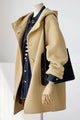 A beige hooded coat displayed on a mannequin, layered over a dark blue denim shirt, with a black shoulder bag hanging from the sleeve.