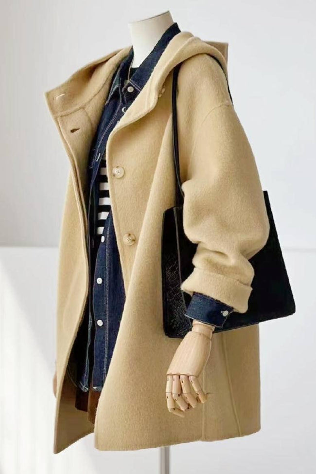 A beige hooded coat displayed on a mannequin, layered over a dark blue denim shirt, with a black shoulder bag hanging from the sleeve.