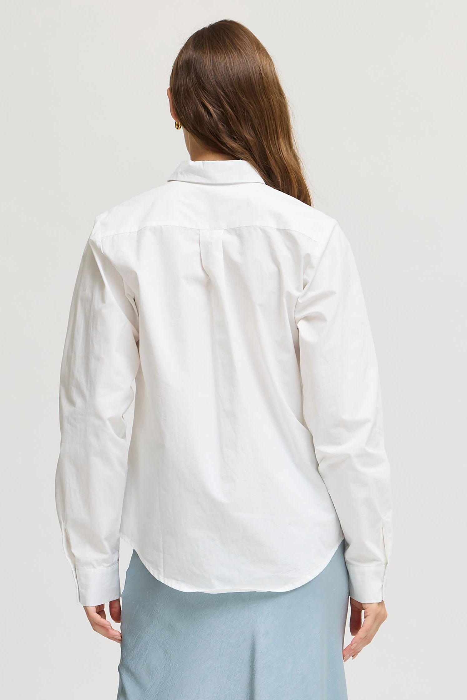 A woman shown from the back wearing a white long-sleeve button-up shirt and a light blue midi skirt, paired with beige high-heeled sandals, against a plain light gray studio background.