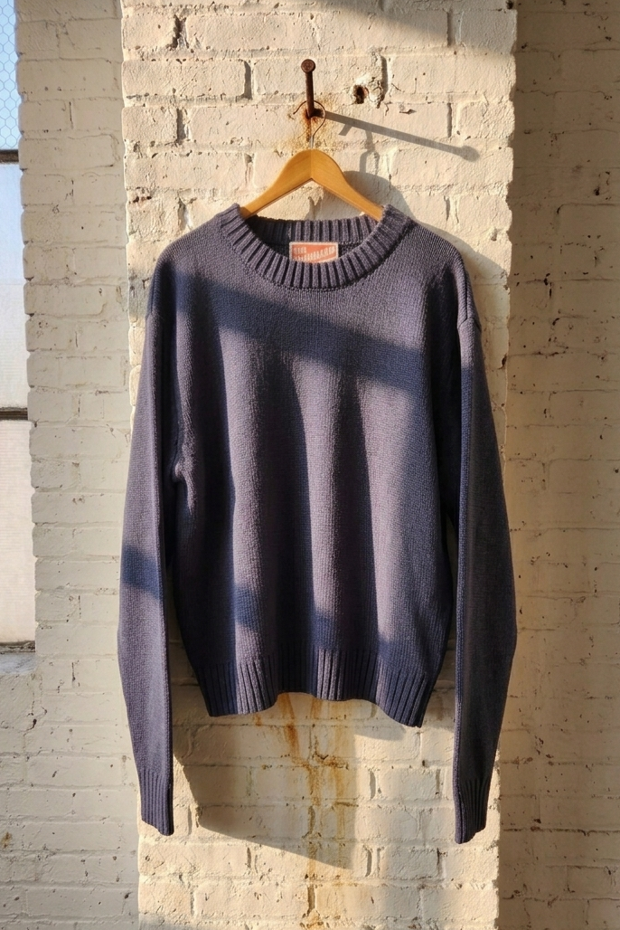 A dark slate blue crewneck knit sweater hanging on a wooden hanger against a white brick wall with warm sunlight and window shadows.