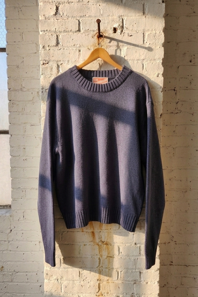 A dark slate blue crewneck knit sweater hanging on a wooden hanger against a white brick wall with warm sunlight and window shadows.