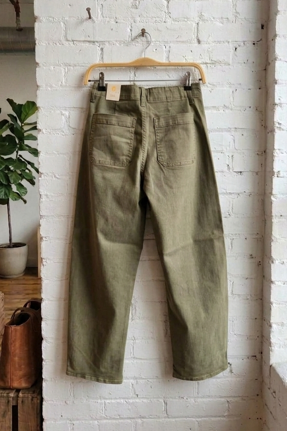 A pair of olive green cropped pants hanging on a wooden hanger against a white brick wall in a loft-style room with a window, wooden crate, and potted plant.