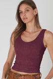 A woman wearing a sleeveless scoop-neck knit top in a deep plum purple color, paired with brown pants and a brown belt, against a light gray studio background.