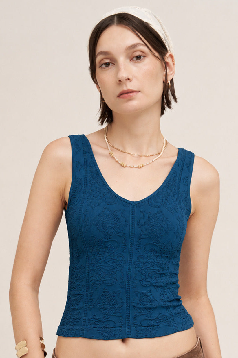 A woman wearing a textured, sleeveless scoop-neck tank top in deep sea blue, paired with brown pants and a white headband, against a light beige background.