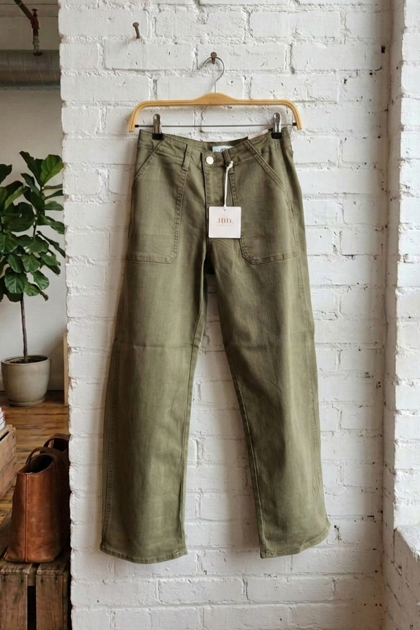 Olive green wide-leg barrel pants hanging on a wooden hanger against a white brick wall in a loft-style room with a window, wooden crate, and potted plant.