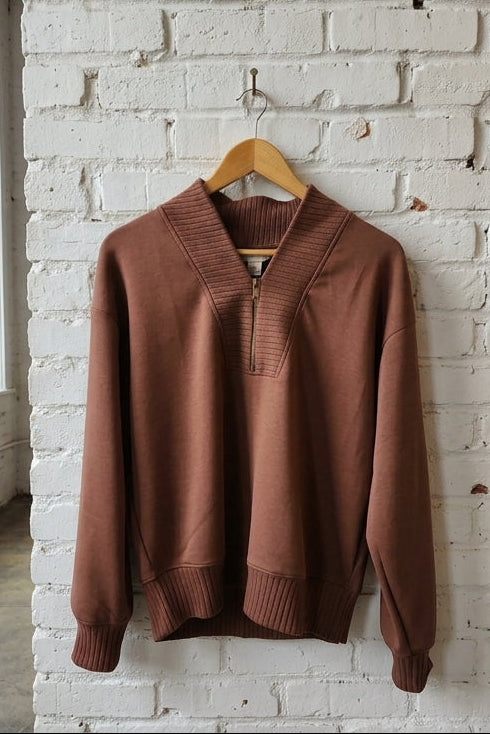 A brown knit sweater with a half-zip V-neck collar hangs on a wooden hanger against a white brick wall in a loft-style room.