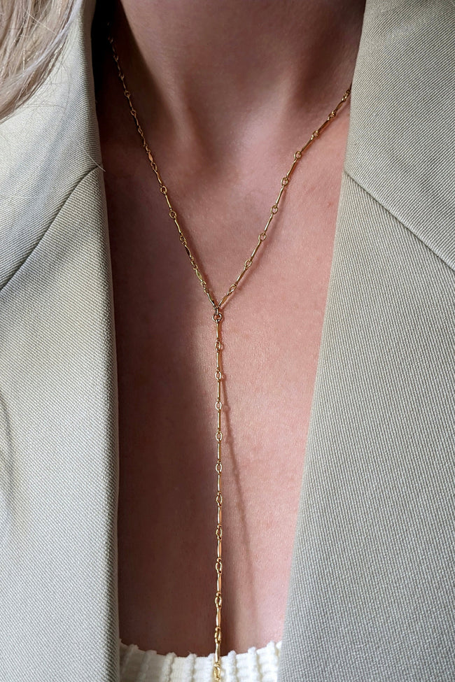 A close-up of a model wearing a delicate gold Y-necklace with a long drop chain over a light beige blazer.