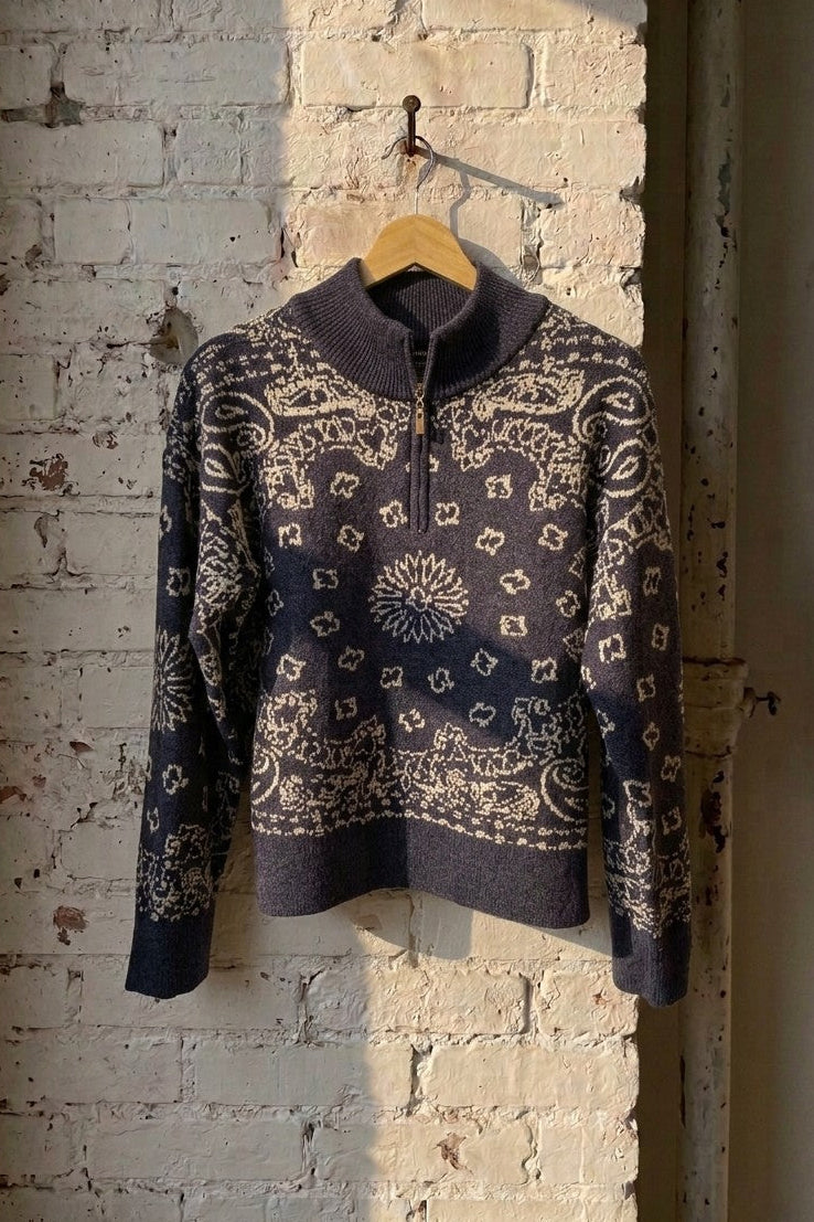 A dark navy and beige bandana patterned knit sweater with a mock neck and half-zip, hanging on a wooden hanger against a distressed white brick wall.