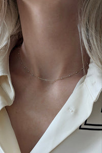 A close-up of a model's neck and collarbone wearing a delicate, thin gold chain necklace with small sparkling stones, paired with a cream-colored collared shirt.