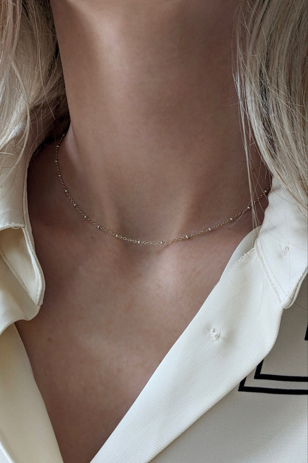 A close-up of a model's neck and collarbone wearing a delicate, thin gold chain necklace with small sparkling stones, paired with a cream-colored collared shirt.